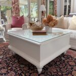 A white square coffee table with a glass top sits on a patterned rug, holding dried flowers, a bowl, and books. Behind it are white sofas with pink and beige pillows in a sunlit living room with large windows.