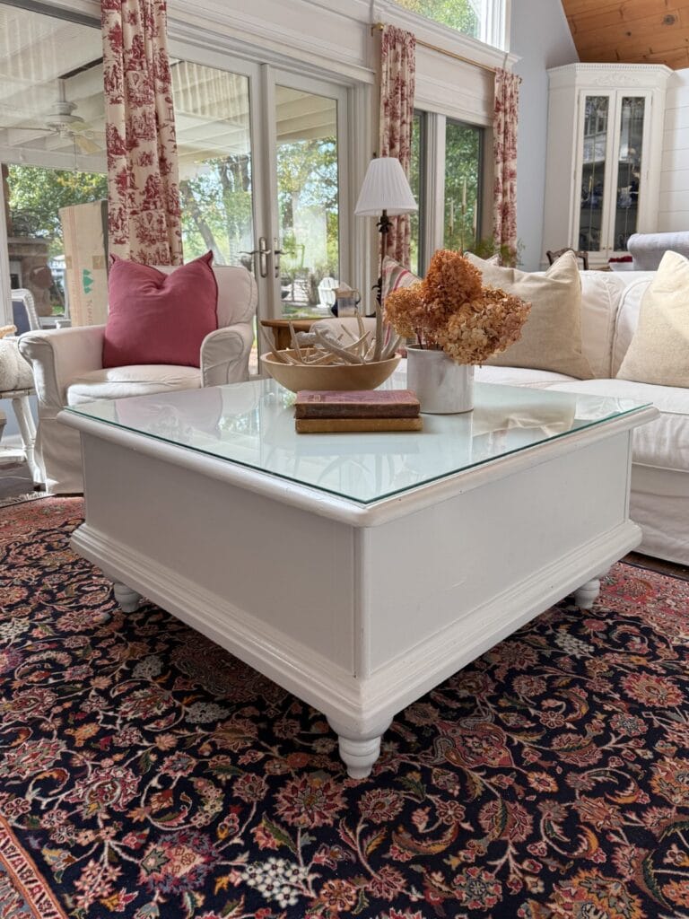 A white square coffee table with a glass top sits on a patterned rug, holding dried flowers, a bowl, and books. Behind it are white sofas with pink and beige pillows in a sunlit living room with large windows.