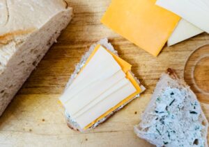 A slice of bread topped with cheddar and Swiss cheese sits on a wooden cutting board, next to another piece of bread with herbed spread and a loaf of bread in the corner.