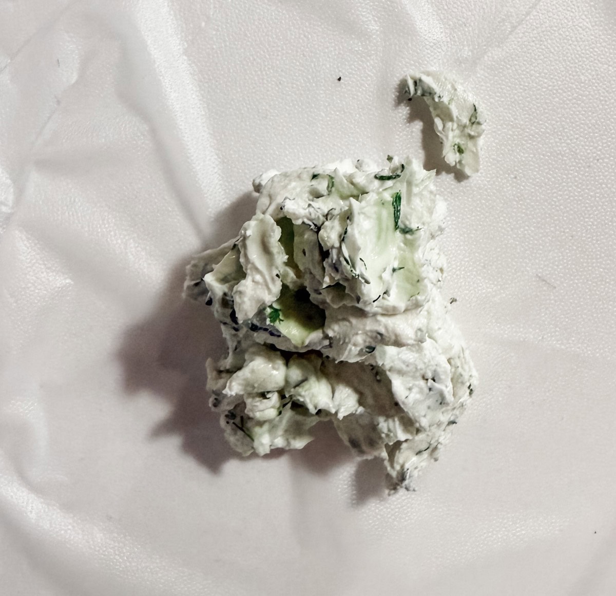A dollop of herbed cream cheese with green herbs is spread on a crinkled white surface.