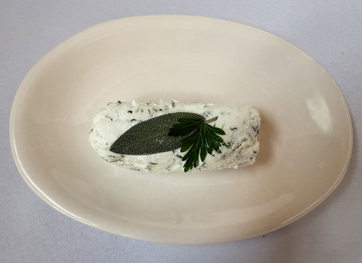 A log of herbed butter garnished with a fresh sage leaf and parsley sits on a plain white oval plate, on a white tablecloth background.