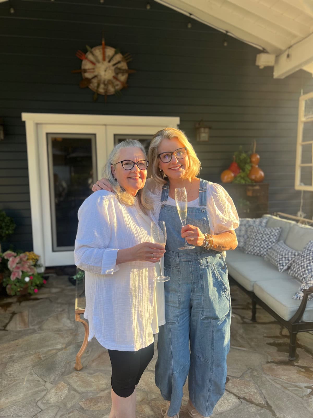Two women smiling and holding champagne glasses stand close together on a sunny patio, showing off their latest style. Behind them is a dark house with a white door, rustic decor, and lush garden plants.