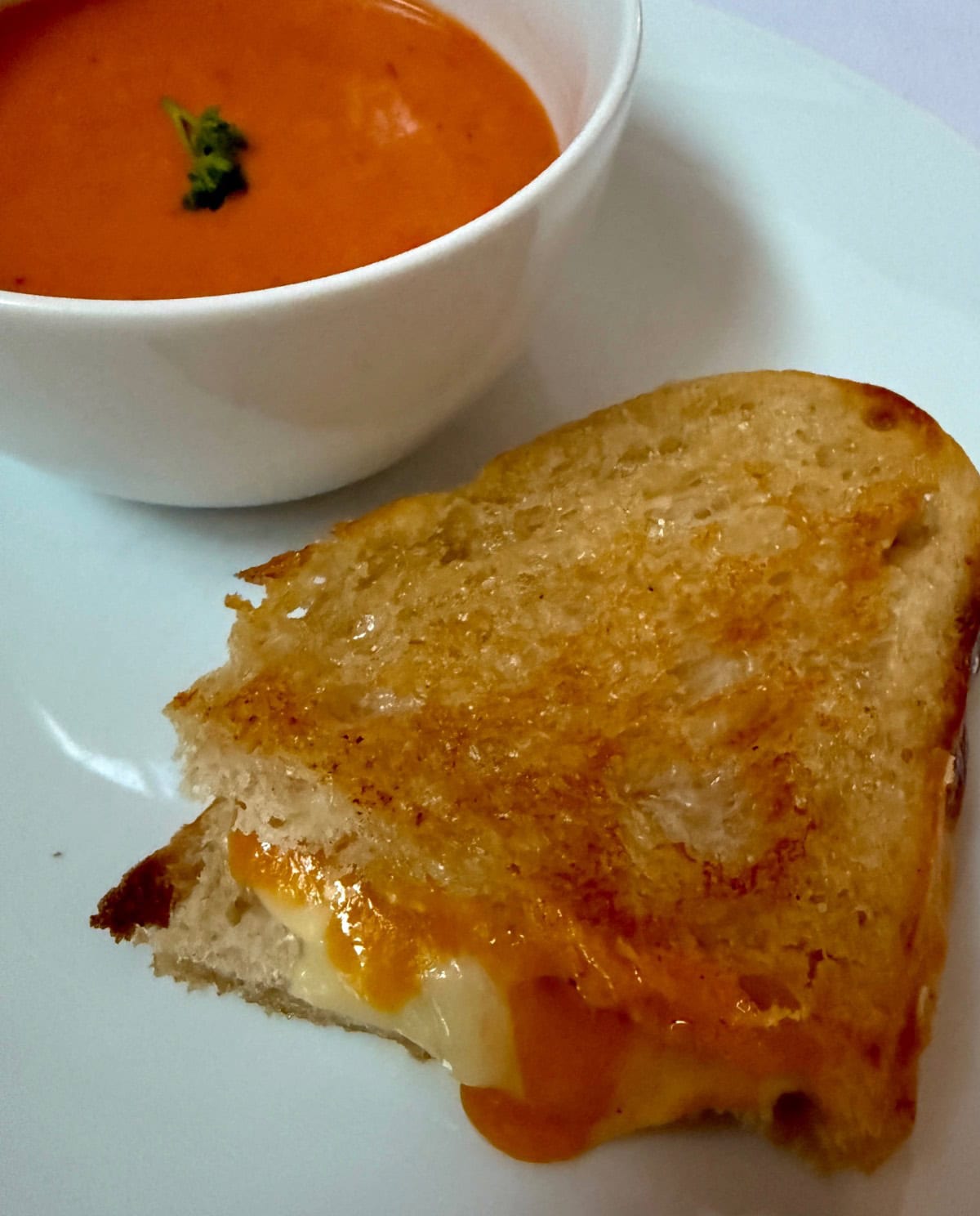 A grilled cheese sandwich with melted cheese oozing out, served on a white plate next to a bowl of creamy tomato soup garnished with a small piece of herb.