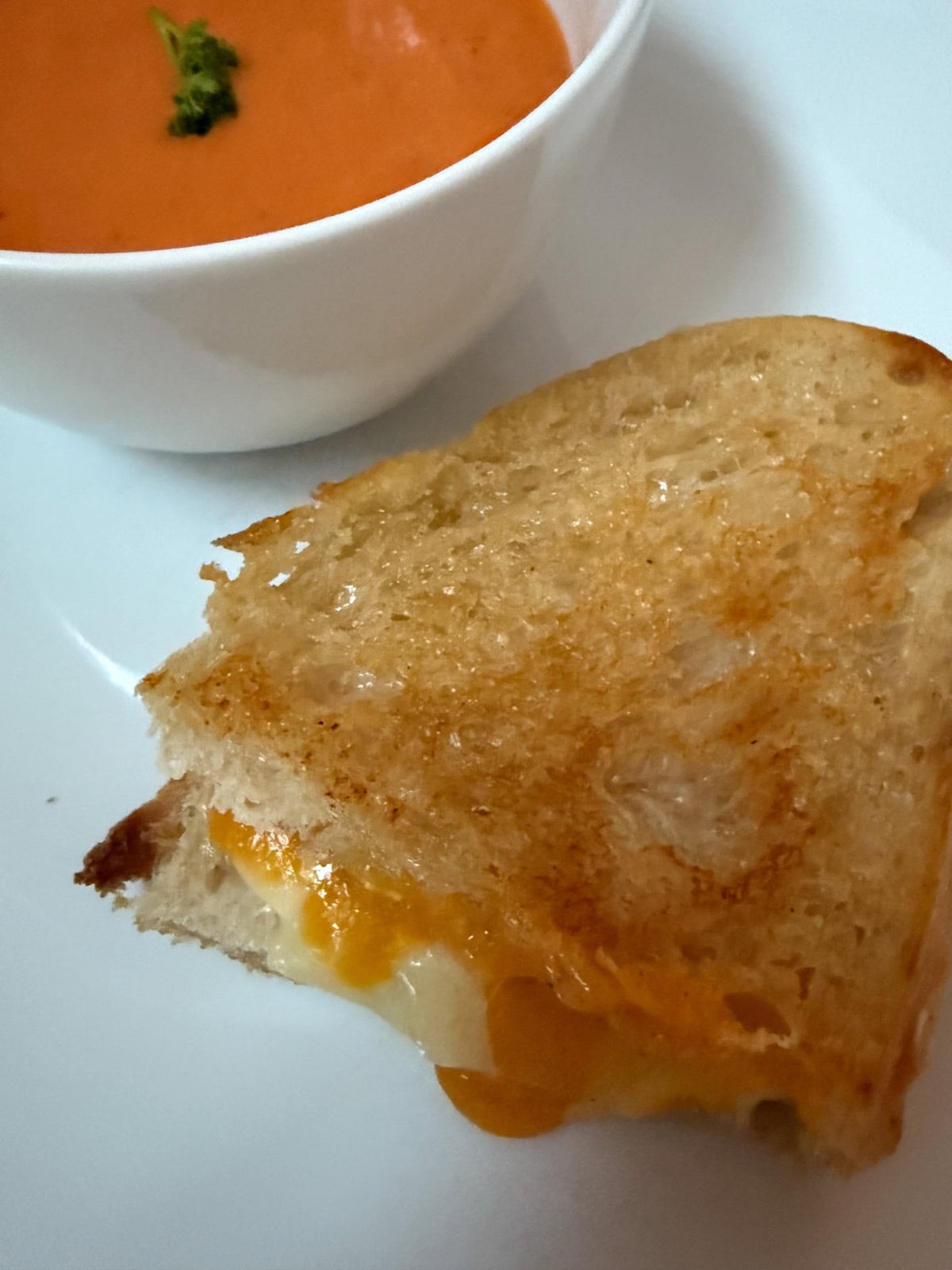 A close-up of a grilled cheese sandwich with melted cheese oozing out, served on a white plate next to a bowl of creamy tomato soup garnished with a small sprig of herbs.