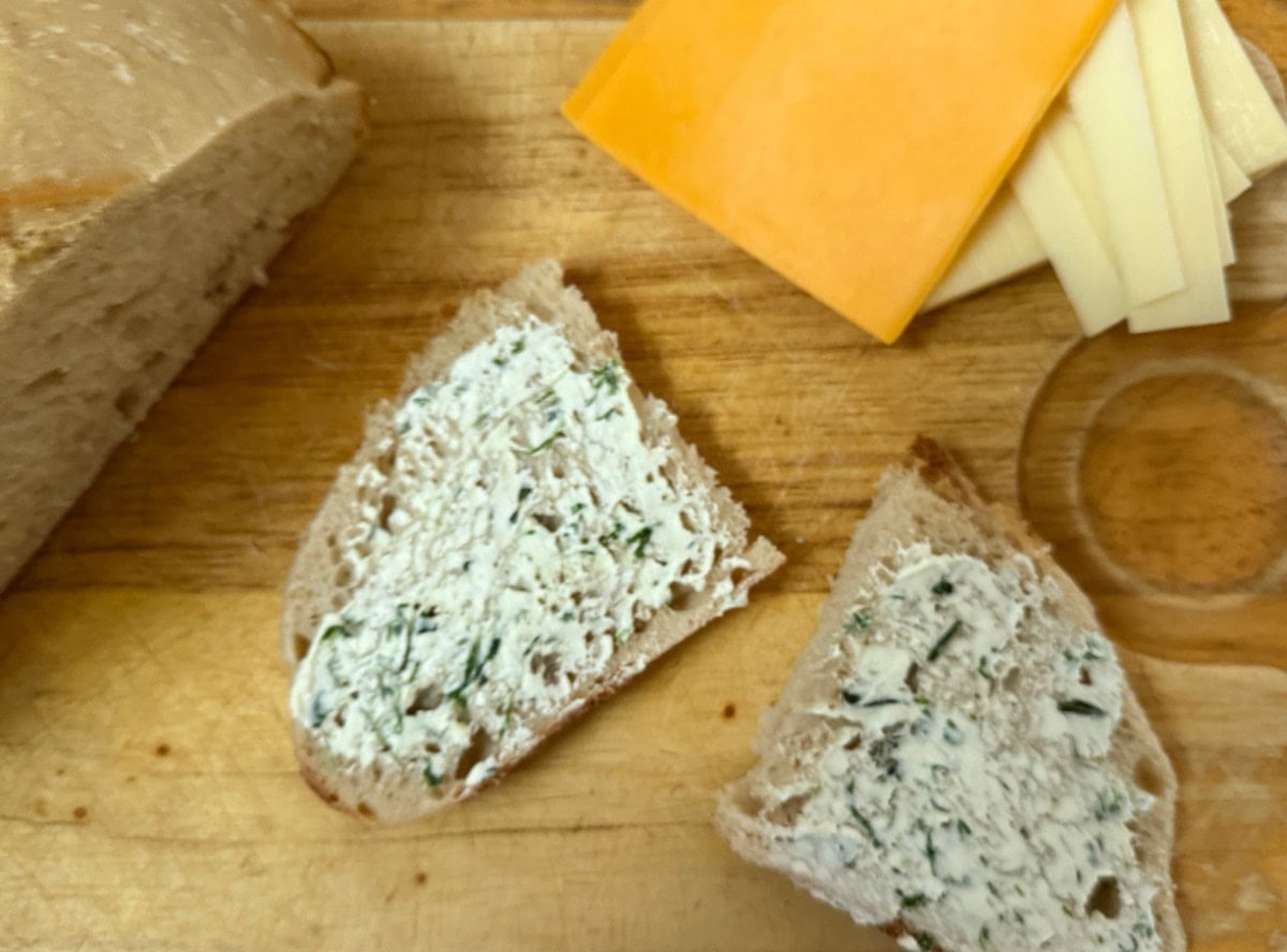 A wooden cutting board with two slices of bread spread with herbed cream cheese, a loaf of bread, and several slices of cheddar and white cheese.