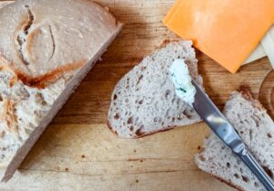 A loaf of rustic bread, two slices on a wooden cutting board, one being spread with herbed cream cheese using a knife; slices of cheddar and Swiss cheese are nearby.