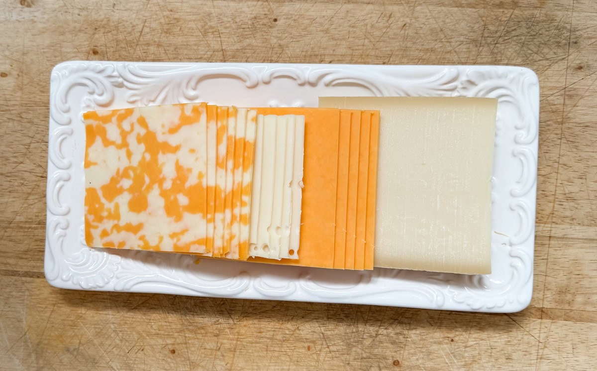 A rectangular white platter with rows of sliced cheeses: marbled orange and white, pale yellow, solid orange, and white slices, displayed on a wooden surface.