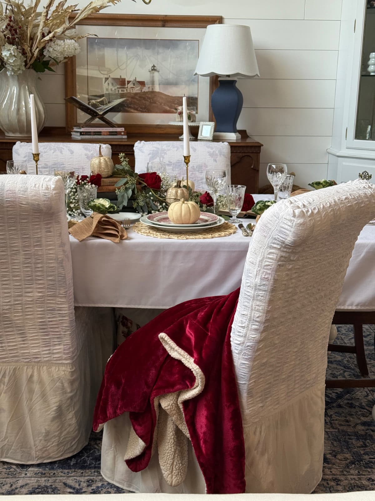 A dining table set for a formal meal with white chairs, gold and book page pumpkins as centerpieces, red roses, candles, and crystal glasses. A red velvet blanket is draped over a chair; a lamp and painting are in the background.