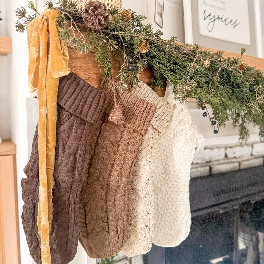 Four knit Christmas stockings in shades of brown and cream hang from a mantel decorated with pine branches, pinecones, and a yellow velvet ribbon above a fireplace.