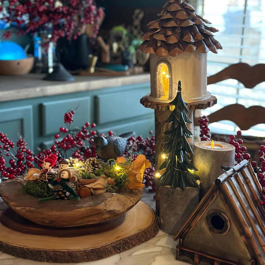 A cozy, rustic holiday display on a marble countertop features a birdhouse, wooden decor, a candle, red berry branches, and small festive figurines with string lights, all set against a kitchen background.