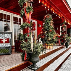 A festive porch decorated for Christmas features greenery, red skis, a Christmas tree in a barrel, string lights, a red rocking chair, and a bench with a cozy blanket on a snowy day.
