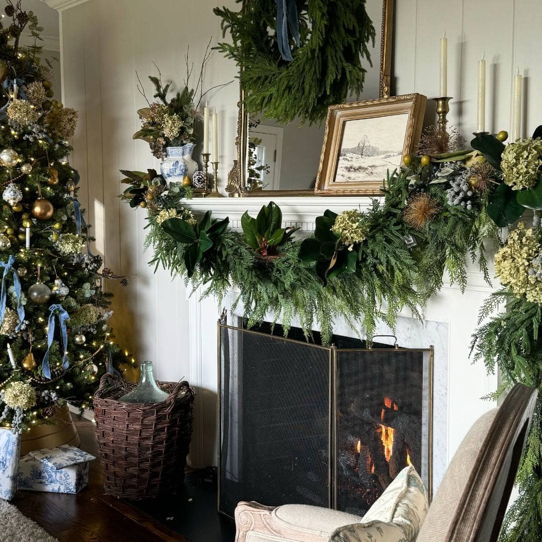 A decorated Christmas tree stands by a fireplace adorned with greenery, hydrangeas, candles, and a framed picture. A fire burns in the fireplace, and a large wreath hangs above the mantle. Presents sit on the floor nearby.