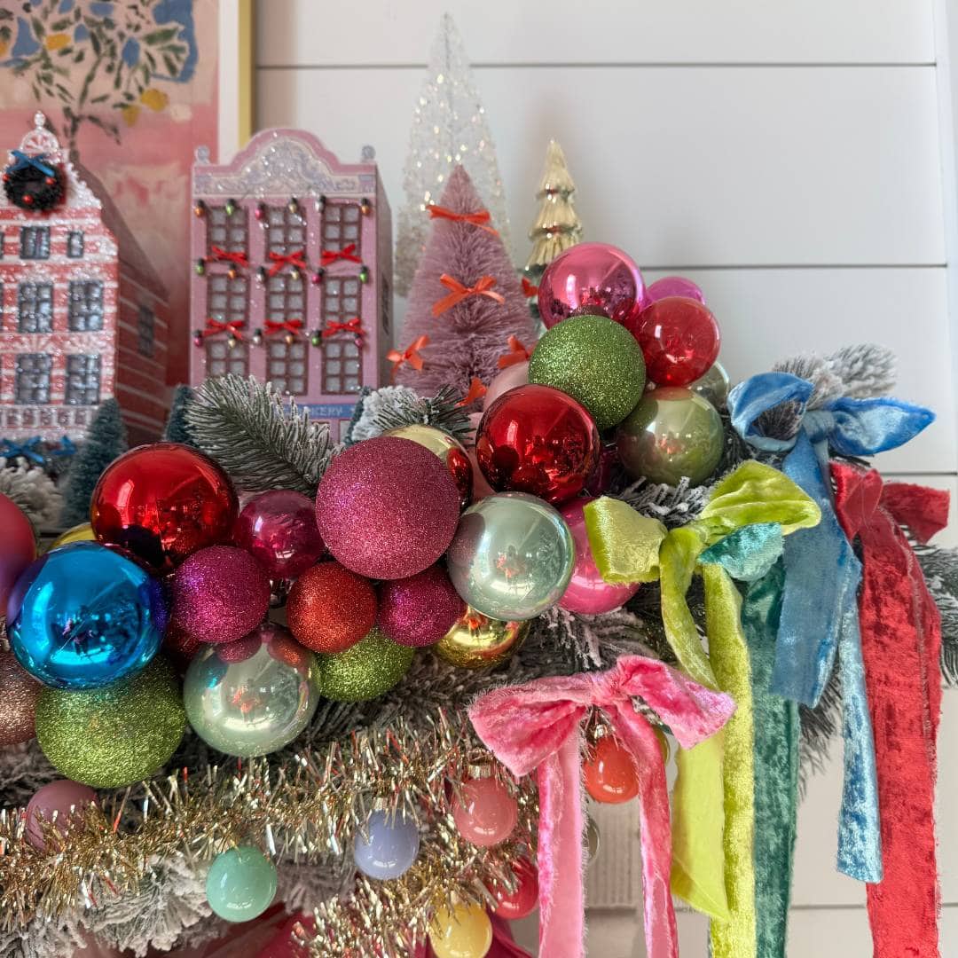 A festive holiday wreath decorated with colorful ornaments, tinsel, and bright velvet bows. Miniature houses and glittery trees are displayed in the background on a white shiplap wall.
