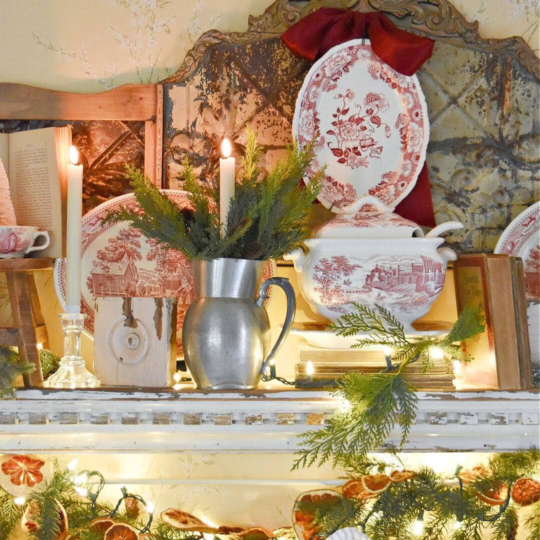 A festive mantel decorated with red and white dishes, a silver pitcher with greenery, candles, old books, and string lights, creating a cozy, vintage holiday atmosphere.