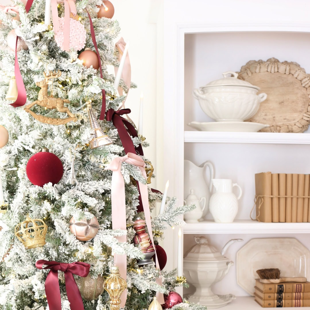 A flocked Christmas tree decorated with pink, gold, and burgundy ribbons and ornaments stands beside a white shelf displaying white dishes, serving ware, and several books with brown covers.