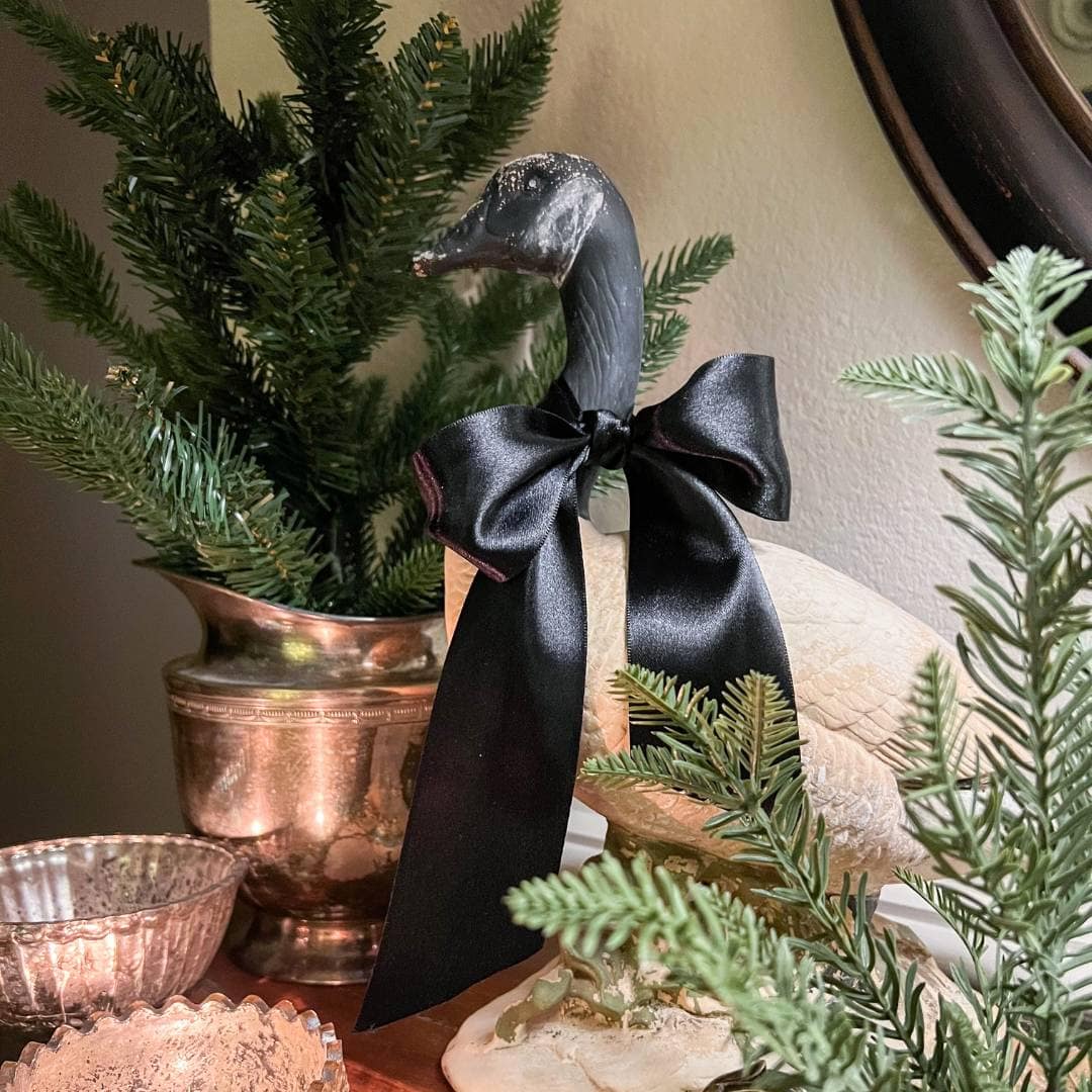A decorative goose statue with a black satin bow around its neck is displayed on a table, surrounded by evergreen branches and silver containers, creating a festive and elegant setting.