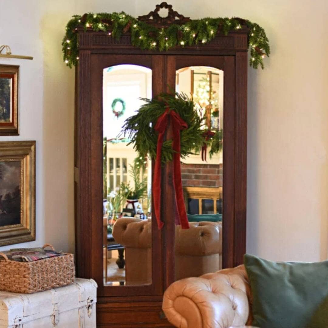 A wooden wardrobe with mirrored doors is decorated for Christmas with a garland of greenery and lights on top and a wreath with a red ribbon hanging on the door. A beige couch with a green pillow sits nearby.