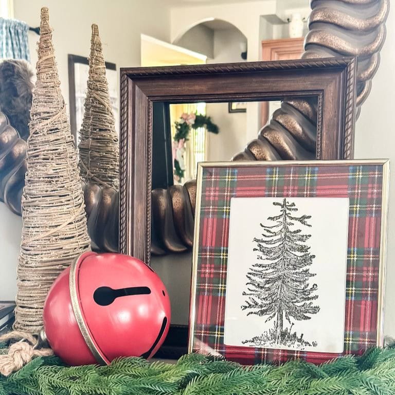 A holiday display with two twine-wrapped trees, a large red jingle bell, and framed art of a pine tree with a red plaid border, set on a surface covered with faux greenery in front of a mirror.