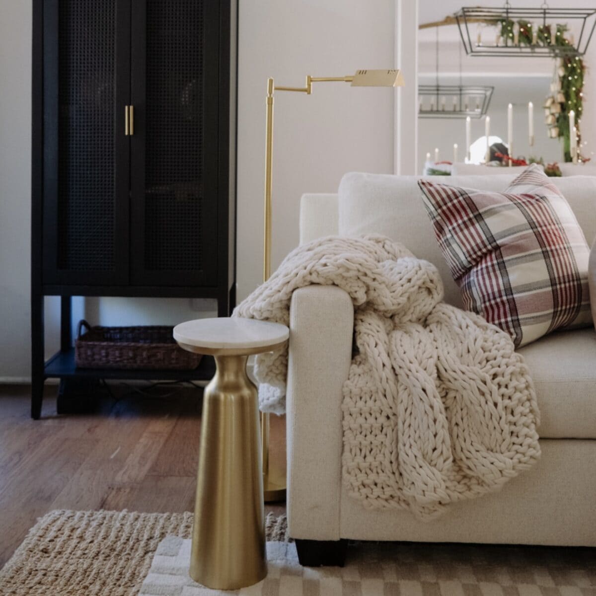 A cozy living room scene with a cream sofa, plaid pillow, chunky knit blanket, gold side table, and gold floor lamp. In the background, a black cabinet and festive holiday decor are visible.