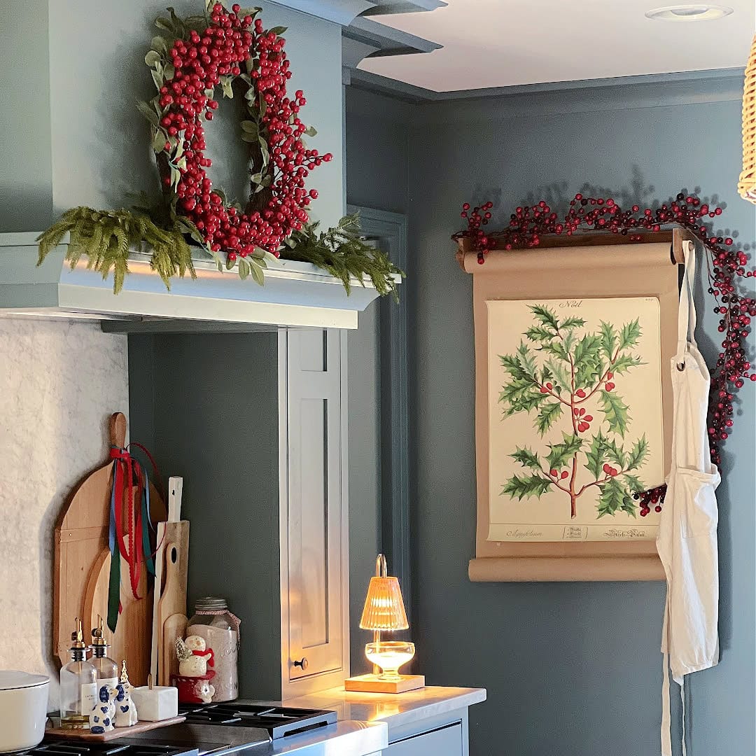 A cozy kitchen with grey-blue cabinets, decorated for the holidays with a red berry wreath and garland. A botanical print hangs on the wall, next to a white apron. Cutting boards and kitchen items rest on the counter.
