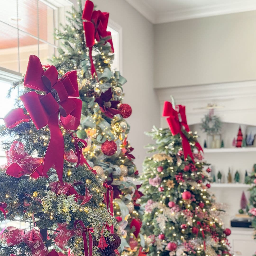Three Christmas trees decorated with red bows, ornaments, and lights stand in a bright living room near a window. Shelves with holiday decorations are in the background.