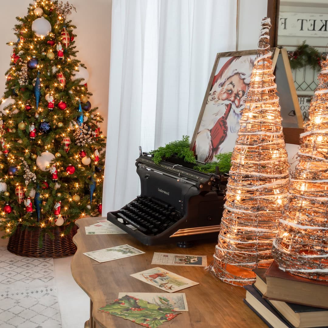A festive room with a decorated Christmas tree, a vintage typewriter on a wooden table, glowing wire Christmas trees, and holiday postcards. A Santa Claus painting and greenery add to the cozy holiday atmosphere.