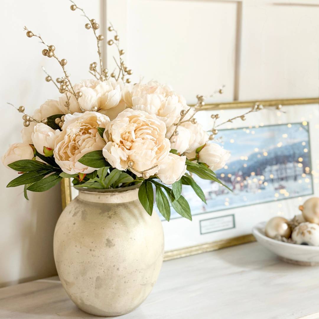 A cream-colored vase filled with pale peony flowers and greenery sits on a light surface beside a framed painting and a bowl of decorative spheres, creating a soft, elegant arrangement.