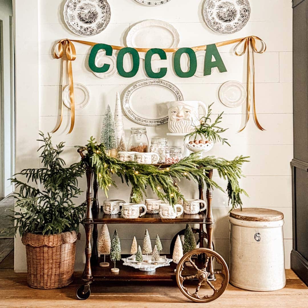 A festive cocoa bar cart decorated with greenery, vintage mugs, jars, ceramic trees, and a "COCOA" banner. Plates are hung on the wall, and a potted evergreen sits to the left of the cart.