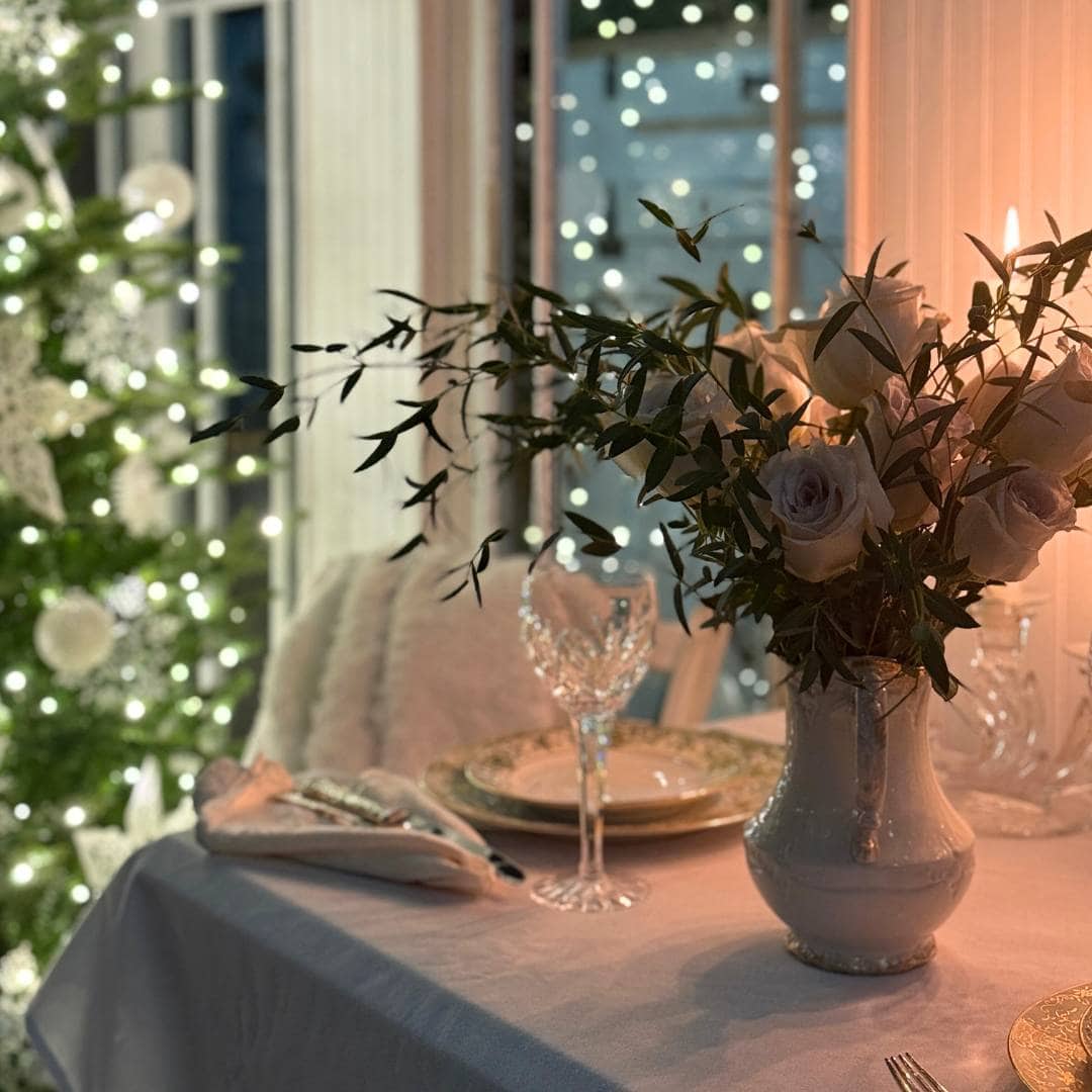 A festive table set with elegant dishes and crystal glasses sits beside a lit Christmas tree. A white vase with flowers and greenery is the centerpiece; warm lights create a cozy holiday atmosphere.