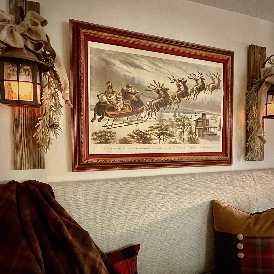 A framed vintage print of Santa in a sleigh pulled by reindeer hangs on a wall, flanked by two rustic lantern sconces with greenery. Cozy plaid blankets and pillows rest on a light upholstered bench below.
