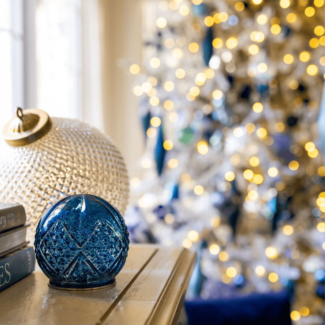 A blue glass ornament and a white textured ornament rest on a table beside stacked books, with a brightly lit, decorated Christmas tree blurred in the background.