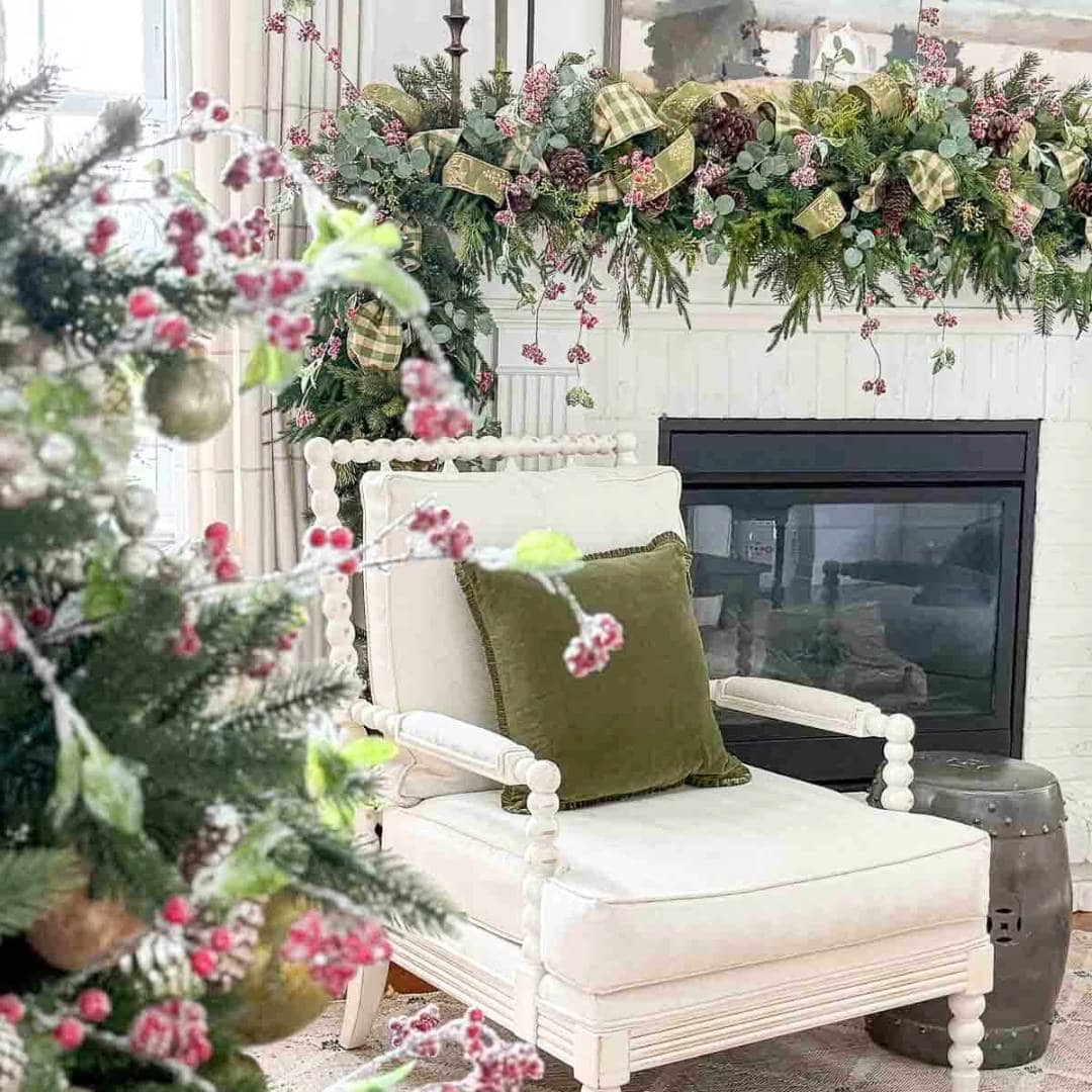 A cozy white armchair with a green cushion sits by a fireplace, which is decorated with a festive garland of greenery, red berries, pinecones, and plaid ribbon. A Christmas tree with ornaments and berries is in the foreground.