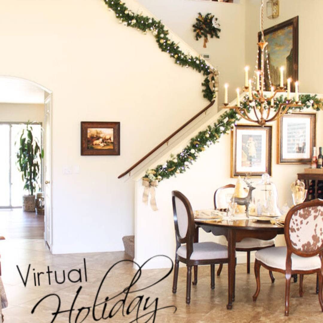 A dining room decorated for the holidays with garland on the staircase railing, a chandelier above a set dining table, framed art on the walls, and sunlight streaming through an open doorway.