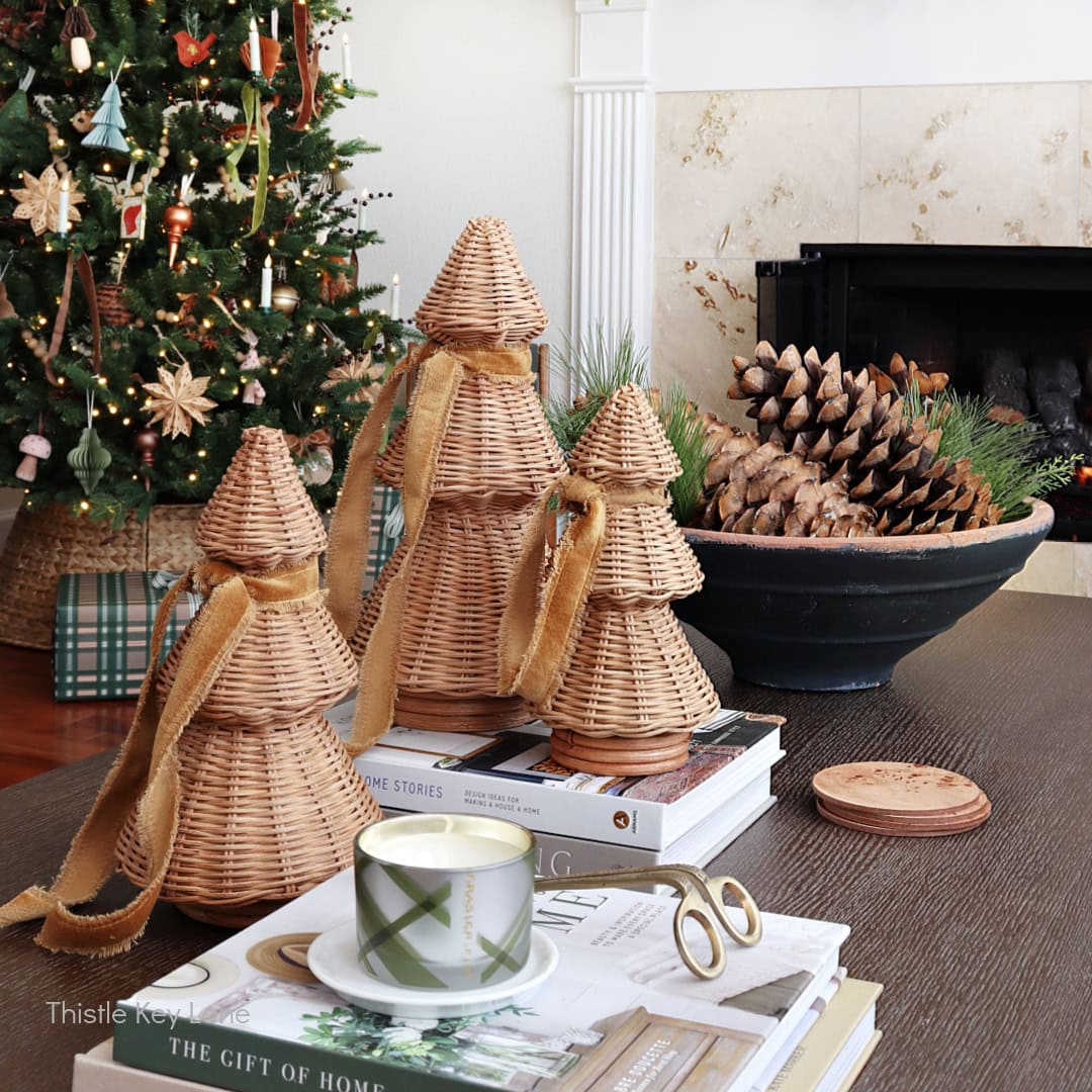 A cozy living room decorated for Christmas with a tree, wicker Christmas tree figures on a coffee table, stacked books, a lit candle, and a bowl of pinecones near a fireplace.
