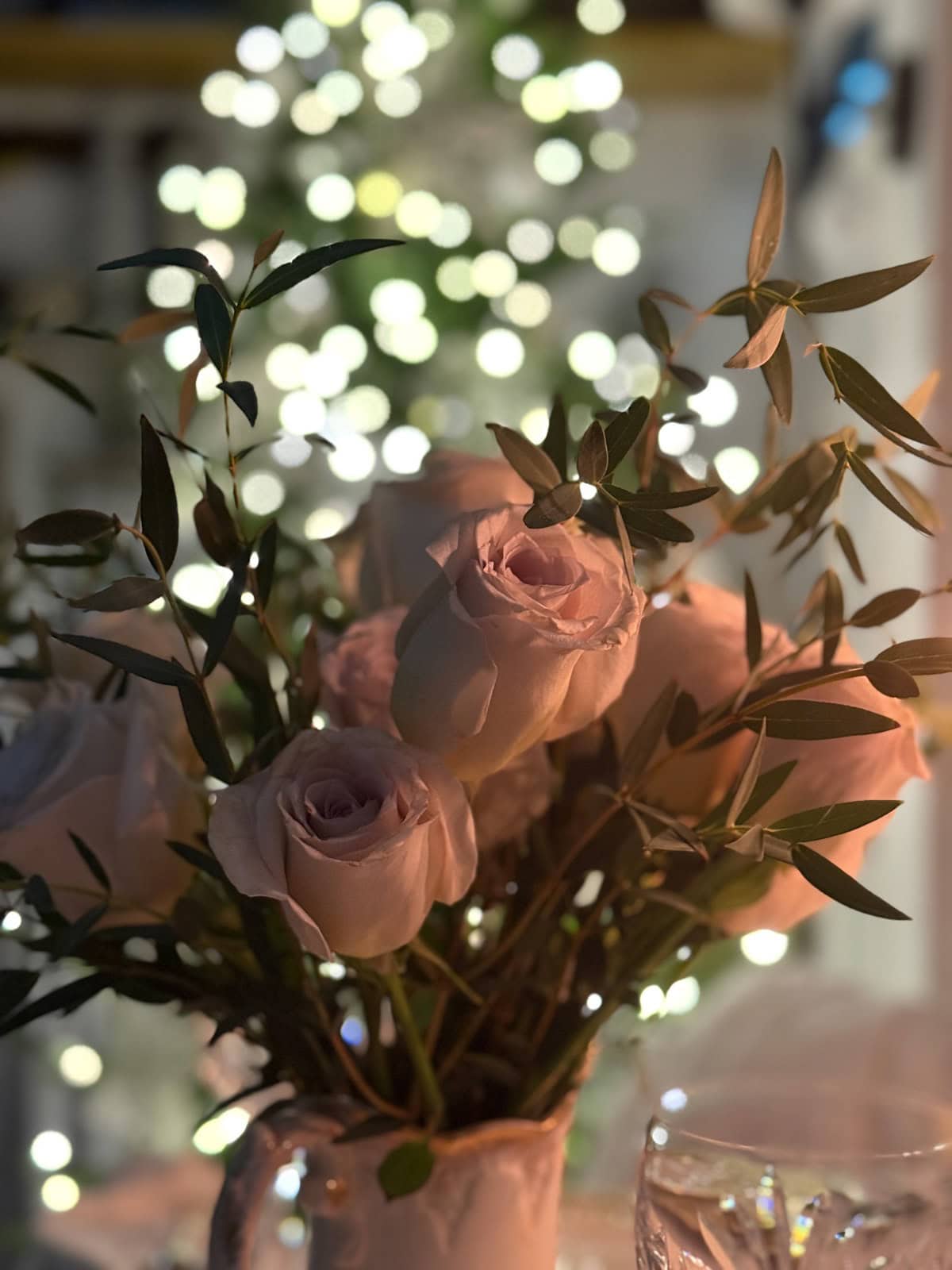 A close-up of a bouquet of pale pink roses with green leaves, softly lit, with blurred twinkling lights and a tree in the background, creating a warm, romantic atmosphere perfect for a holiday home tour.