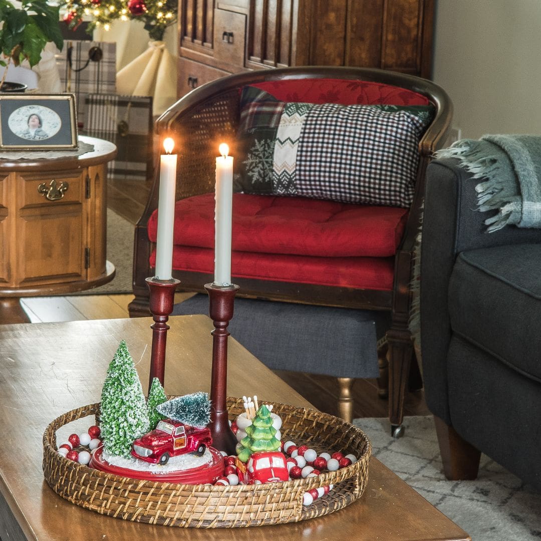 A cozy living room decorated for Christmas with a wicker tray on a coffee table holding two lit candles, small festive ornaments, and mini trees. A red-cushioned chair and holiday decor are in the background.