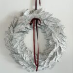 A wreath made from vintage hymnal music, shaped like leaves, hangs on a white wall. It is decorated with a maroon ribbon tied in a bow at the top, with elegant long tails draping down.