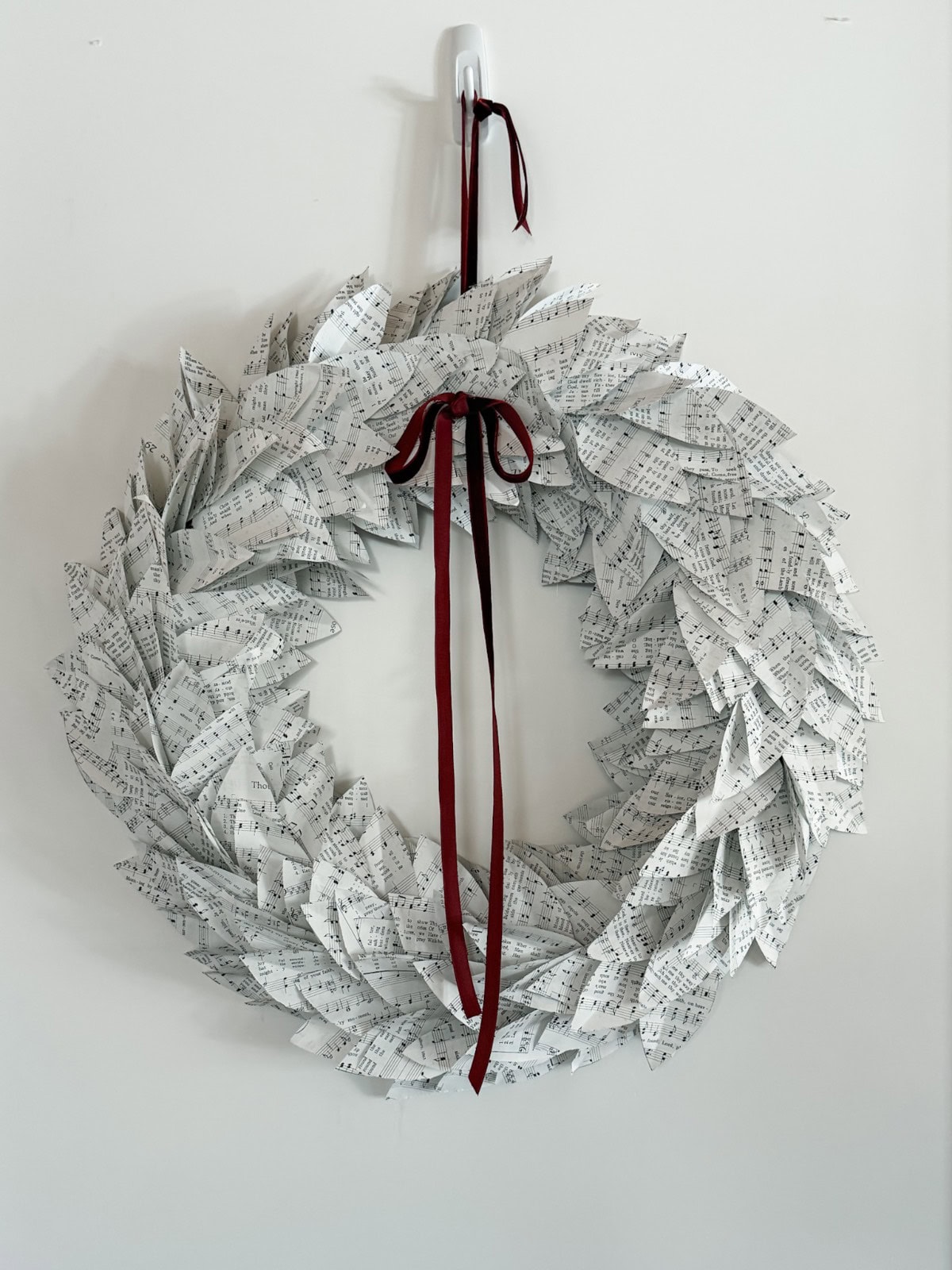 A wreath made from vintage hymnal music, shaped like leaves, hangs on a white wall. It is decorated with a maroon ribbon tied in a bow at the top, with elegant long tails draping down.