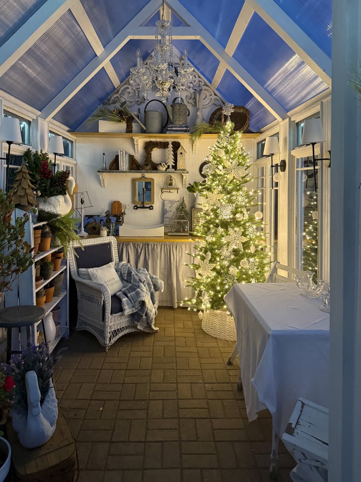 A cozy greenhouse-inspired sunroom, elegantly decorated for Christmas, features a lit tree, white wicker chair with plaid blanket, shelves of plants and decor, and a chandelier hanging from a blue-paneled ceiling—perfect for your holiday home tour.