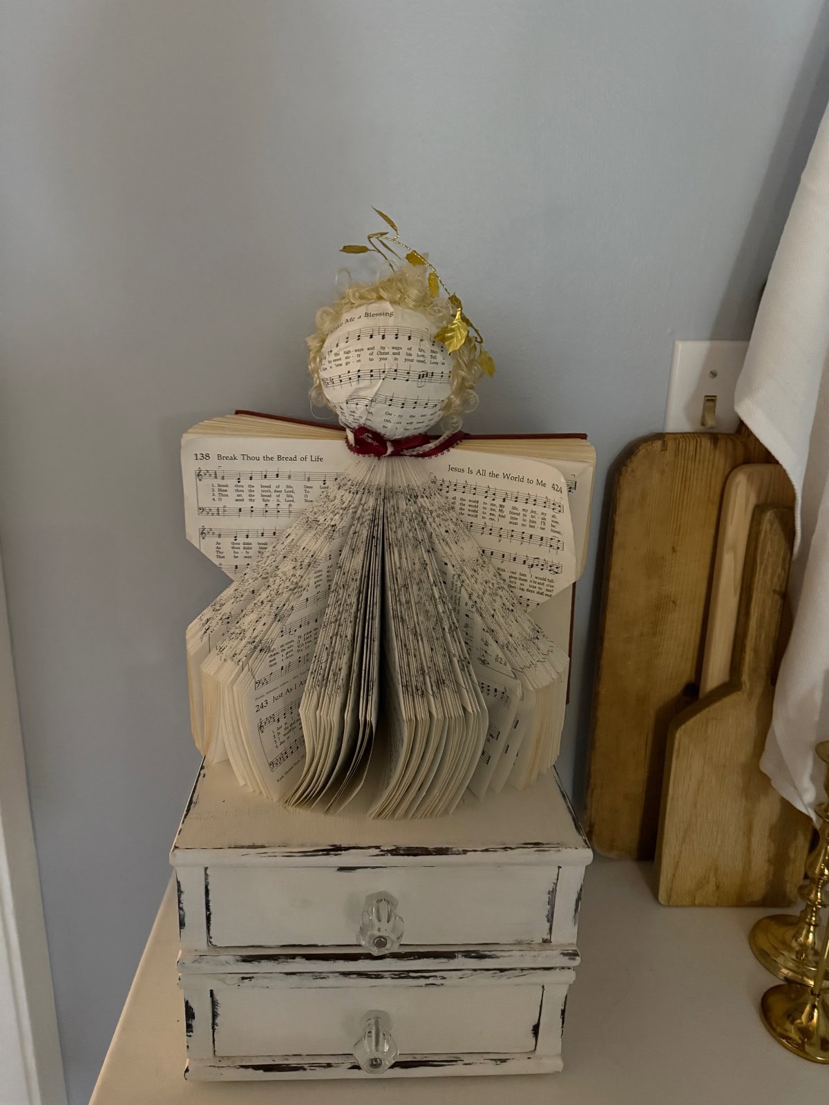 A handmade angel figurine crafted from folded book pages, reminiscent of a wreath made from vintage hymnal music, is decorated with a ribbon and golden halo atop a distressed white jewelry box, with wooden boards in the background.