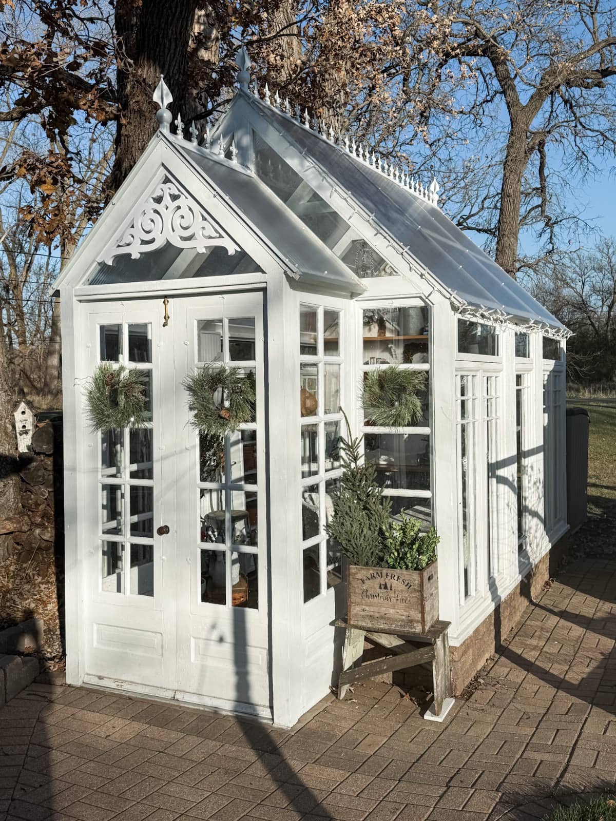 A cozy greenhouse with ornate trim and glass panels stands on a brick patio, adorned for Christmas. Twin wreaths hang on double doors, and potted plants fill a wooden crate—perfect for a Holiday Home Tour amid trees and blue sky.