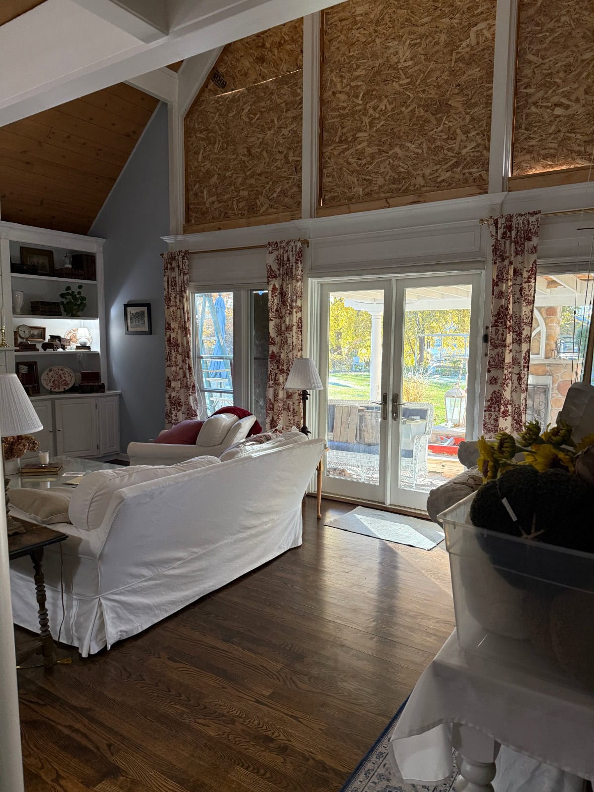 A cozy living room with white sofas, floral curtains, and large windows letting in sunlight. Built-in shelves and wooden floors blend with the latest trend of exposed unfinished upper walls near the ceiling.