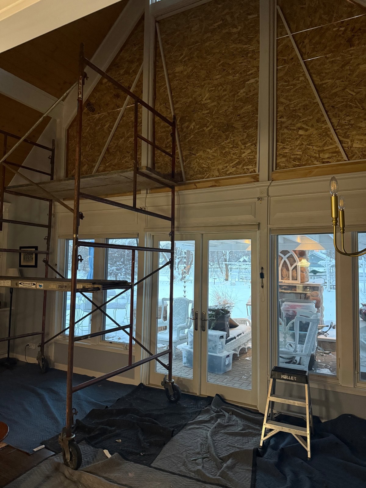 A tall indoor scaffold stands in front of large windows and doors, with the upper wall covered in plywood for the latest renovations. Blankets cover the floor, and snow is visible outside the windows.