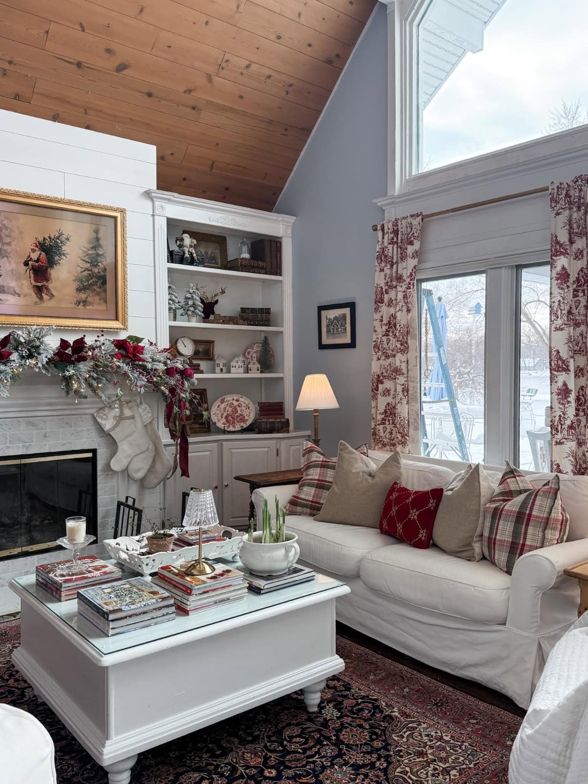 A cozy living room with a white sofa, plaid and red pillows, a white coffee table with books, the latest holiday decor on the fireplace with stockings, shelves with decor, and large windows showing a snowy outdoor scene.