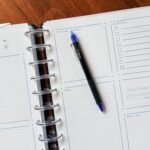 A 2026 Planner open to a weekly spread with sections for priorities, notes, tasks, and lists. A blue and black pen rests diagonally on the page. The background is a wooden table.
