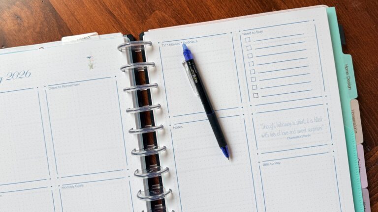 A 2026 Planner open to a weekly spread with sections for priorities, notes, tasks, and lists. A blue and black pen rests diagonally on the page. The background is a wooden table.