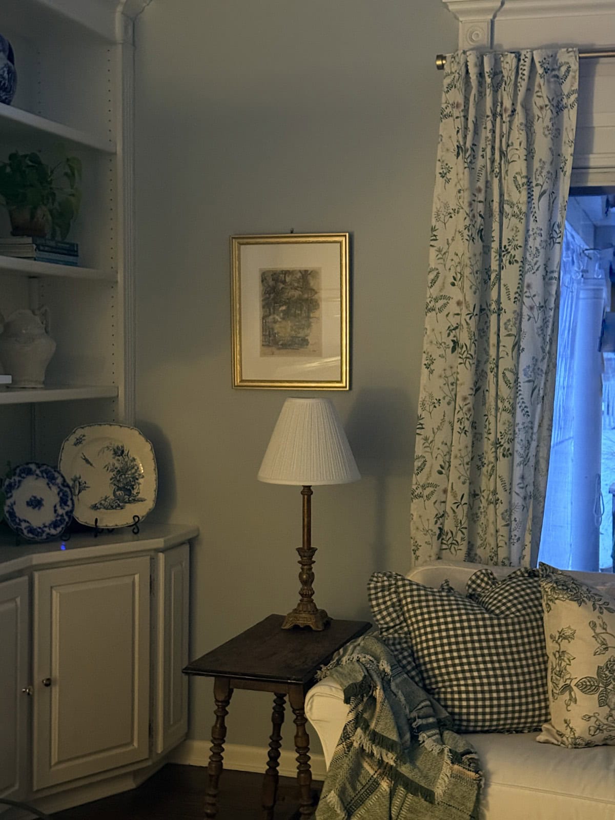 A cozy living room corner evokes a calm winter home with a white couch, green and white patterned pillows, a plaid blanket, wooden side table and brass lamp, floral curtains, framed picture, and blue-and-white decorative plates.