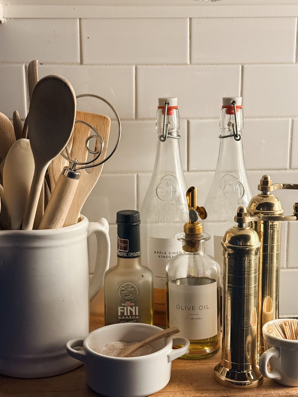 A kitchen countertop in a calm winter home features wooden utensils in a white ceramic holder, bottles of olive oil and vinegar, a salt bowl, and grinders, all set against a classic white subway tile backsplash.