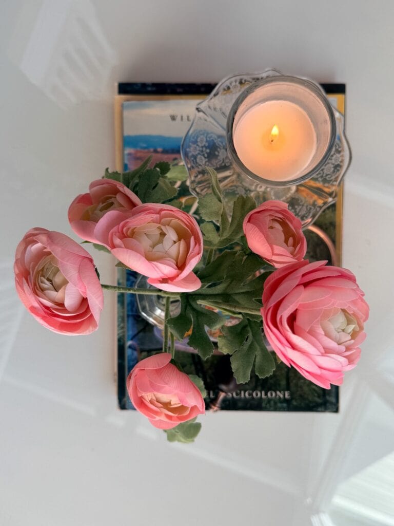 A top-down view of pink ranunculus flowers in a vase, placed next to a lit candle in a glass holder, all resting on two stacked books—perfect for creating a calm winter home on a white surface.