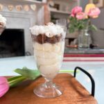 A layered dessert in a glass sits on a wooden tray, featuring rich chocolate mousse and airy mascarpone cream. Pink tulips and stacked books decorate the cozy, light-filled living room in the background.